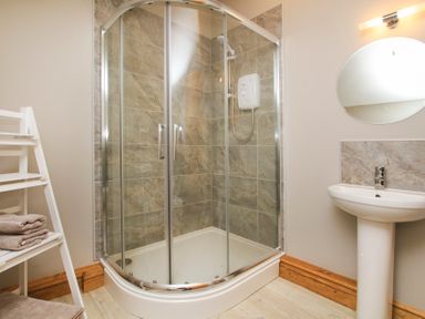 A bathroom with a corner shower enclosure a white pedestal sink a round mirror and a white shelf with towels at Mulberry Cottage in Pentraeth