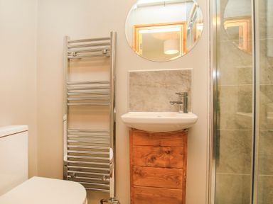 A bathroom with a toilet sink with wooden cabinet round mirror heated towel rail and glass shower door at Partridge Cottage in Menai Bridge