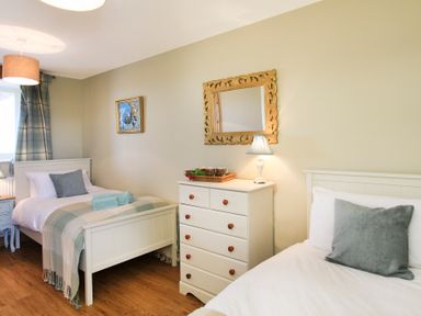 A bedroom with two single beds a white chest of drawers a wall mirror and a window with curtains at Teal Cottage in Menai Bridge