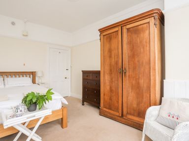 A bedroom with a wooden bed white bedding a wooden wardrobe a wooden chest of drawers a white chair with a cushion and a small table with a plant and teapot at Windygates in Rhosneigr