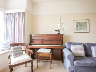 A living room with a wooden piano a wooden chair a wooden piano stool a blue couch with cushions and striped curtains at Windygates in Rhosneigr