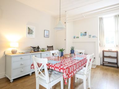 A dining room with a red patterned tablecloth on a wooden table surrounded by white chairs and a light blue sideboard with a lamp at Windygates in Rhosneigr