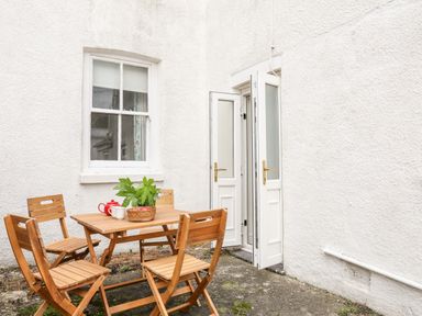 An outdoor patio area with a wooden table and four chairs next to a white wall with a window and an open door at Windygates in Rhosneigr