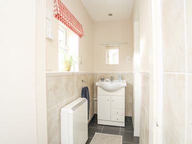 A bathroom with a white sink cabinet a small mirror a radiator and a window with a red and white checked blind at Windygates in Rhosneigr