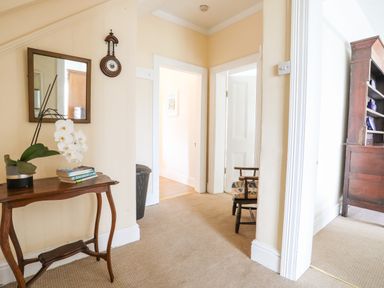 A hallway with a wooden table holding a plant and books a wall mirror a clock two open doors showing a chair and a wooden shelf at Windygates in Rhosneigr