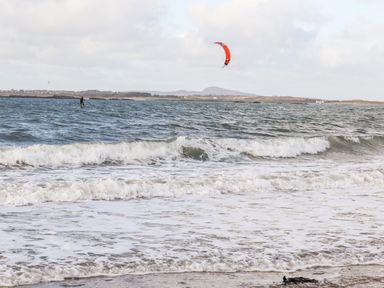 A beach with waves and a person kite surfing on the water with hills in the background at Windygates in Rhosneigr