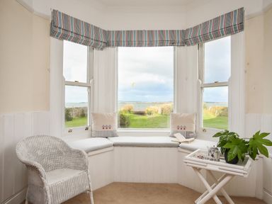 A seating nook with a white wicker chair cushions on a window bench a small table with a plant and tea set and a view of the sea at Windygates in Rhosneigr
