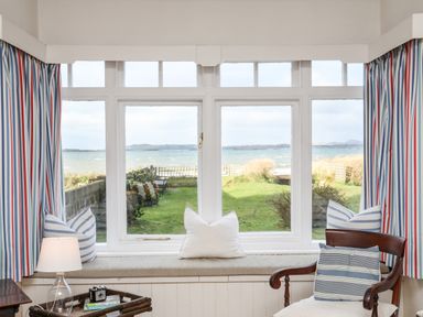 A window seat with cushions and a view of a garden and water with curtains and a chair at Windygates in Rhosneigr