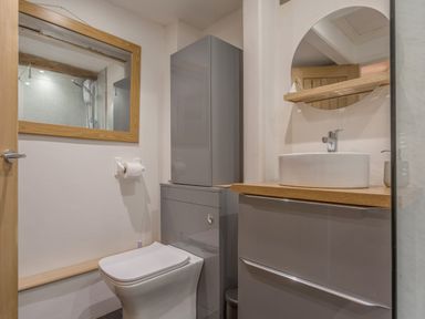 A bathroom with a toilet, grey cabinets, a round mirror above a sink and a wooden shelf at Potters by the Sea in Sennen