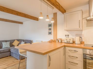 A kitchen area with wooden countertop and white cabinets next to a living space with a gray sofa and wooden beams on the ceiling at Potters by the Sea in Sennen