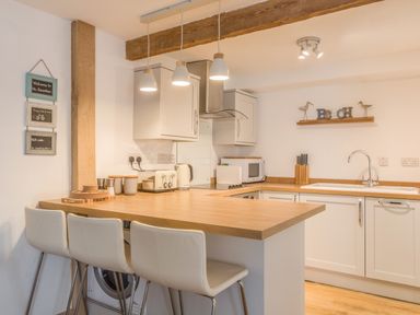 A kitchen with a wooden countertop island with three white chairs a toaster kettle microwave knives and a sink at Potters by the Sea in Sennen
