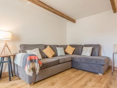 A corner grey sofa with pillows and a blanket next to a lamp on a wooden floor at Potters by the Sea in Sennen