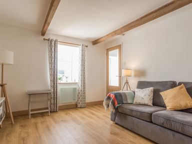 A living room with a gray sofa with pillows and a blanket a window with patterned curtains a wooden floor a floor lamp a small table and a wooden beam ceiling at Potters by the Sea in Sennen