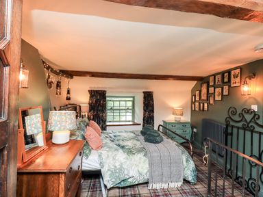 A bedroom with a bed, dresser with mirror and lamp, small cabinet, window with curtains and framed pictures on the wall at Bonny Bits Cottage in Garrigill near Alston