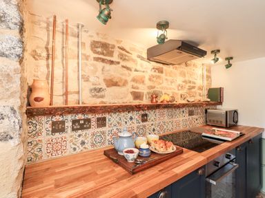 A kitchen countertop with a wooden surface an electric stovetop a toaster a tray with a teapot cups and scones against a tiled and stone wall at Bonny Bits Cottage in Garrigill near Alston