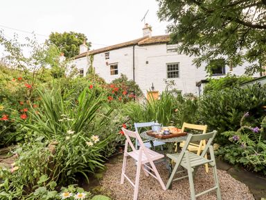 An outdoor garden with a round table set with tea and chairs in front of a white cottage at Bonny Bits Cottage in Garrigill near Alston