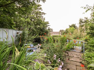 A garden with a stone path, metal gate, flowering plants, and a table with chairs at Bonny Bits Cottage in Garrigill near Alston