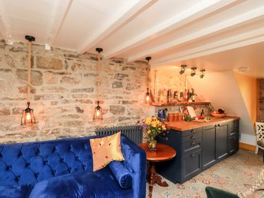 A room with a blue couch a wooden side table flowers hanging pendant lights and a kitchen counter with bottles and fruit at Bonny Bits Cottage Garrigill near Alston