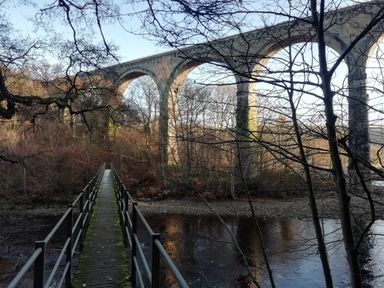 A narrow footbridge over a river with a stone railway viaduct and trees at Bonny Bits Cottage in Garrigill near Alston