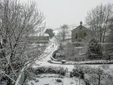 A snow-covered rural road with trees fields and stone buildings at Bonny Bits Cottage in Garrigill near Alston