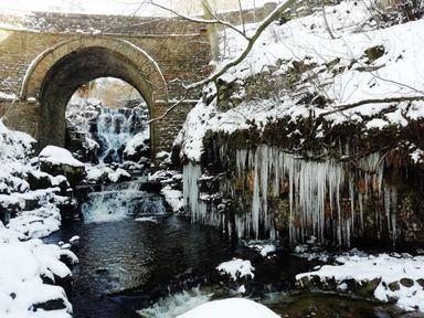 A stone arch bridge over a flowing creek with waterfall and icicles on rocks and snow at Bonny Bits Cottage in Garrigill near Alston