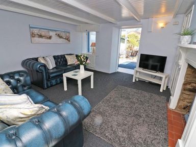 A living room with blue leather sofas a white table flowers on it a TV and an open door to the outside at Pebble Cottage in Portreath