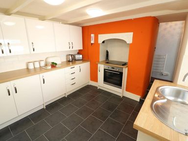 A kitchen with white cabinets an orange accent wall built in oven and black tile floor at Pebble Cottage in Portreath