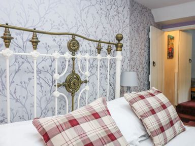 A bedroom with a white and brass bed frame red and white plaid cushions wallpaper with a branch pattern and a bedside lamp at Pebble Cottage in Portreath