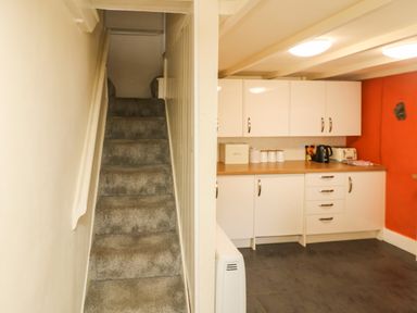 A staircase with carpet leading up next to a kitchen with white cabinets on a red wall at Pebble Cottage in Portreath