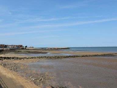 A beach with rocks and low tide water near a coastline with buildings and wind turbines in the distance at Sea Breeze in Rhos-on-Sea