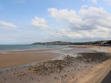 A beach with rocks and sand near a shoreline with buildings and hills in the background at Sea Breeze in Rhos-on-Sea