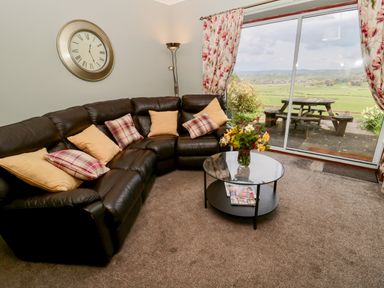A living room with a black leather corner sofa and cushions a glass coffee table and sliding glass doors opening to a patio at Forge Cottage in Rothbury