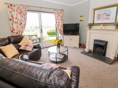 A living room with leather sofas a round glass coffee table a television on a stand a fireplace and sliding glass doors with floral curtains at Forge Cottage in Rothbury