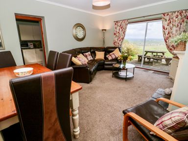 A living room with a brown leather sectional sofa and a wooden dining table with chairs next to large sliding glass doors with floral curtains at Forge Cottage in Rothbury
