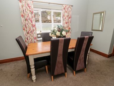 A dining room with a wooden table surrounded by six high backed chairs and floral curtains at Forge Cottage in Rothbury