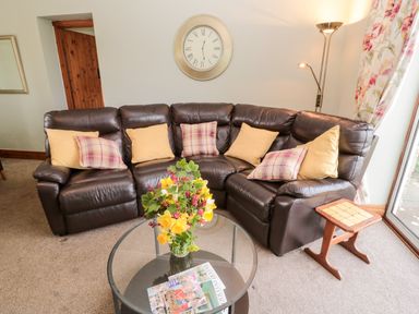 A living room with a brown leather corner sofa with cushions a glass coffee table with flowers and magazines and a wooden side table at Forge Cottage in Rothbury