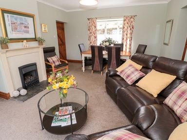 A living room with a leather sofa with cushions a glass coffee table with flowers a fireplace and a dining table with chairs at Forge Cottage in Rothbury