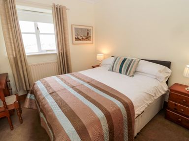 A bedroom with a double bed striped bedspread pillows two bedside tables with lamps a window with curtains and framed artwork on the wall at Forge Cottage in Rothbury