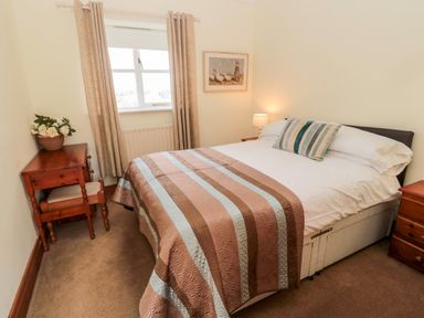 A bedroom with a bed covered with a striped blanket and pillows a bedside table with a lamp a wooden desk with a stool and a flower vase at Forge Cottage in Rothbury
