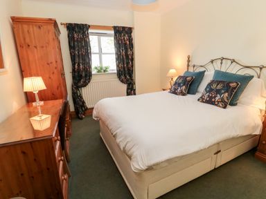 A bedroom with a bed a wooden dresser wooden wardrobe two bedside tables lamps window with curtains and a green carpet at Forge Cottage in Rothbury
