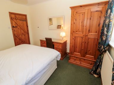 A bedroom with a bed a wooden wardrobe desk with drawers and a chair a wooden door and a window with patterned curtains at Forge Cottage in Rothbury
