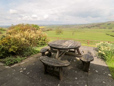 An outdoor patio area with a round wooden picnic table with benches overlooking green fields with a wooden fence and hills in the distance at Forge Cottage in Rothbury