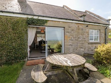 A stone house with a sliding glass door and a round wooden picnic table outside at Forge Cottage in Rothbury