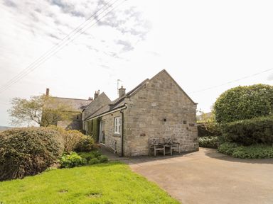 Stone house with two wooden chairs outside surrounded by bushes and grass at Forge Cottage in Rothbury