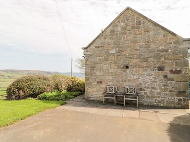 An exterior stone wall with two wooden chairs on a paved area next to bushes and a grassy field at Forge Cottage in Rothbury