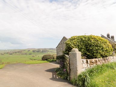 A driveway with a stone wall and bushes next to a stone building overlooking green fields and hills at Forge Cottage in Rothbury