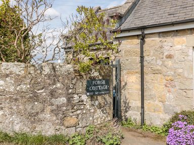 An entrance gate with a stone wall and a sign reading Forge Cottage attached to a stone building at Forge Cottage in Rothbury