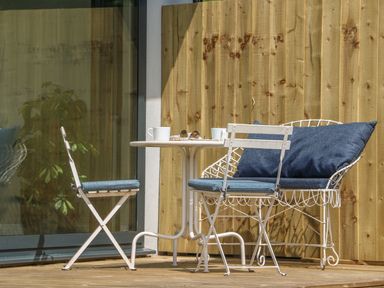 A patio with a small round table and three white metal chairs with blue cushions next to a wooden fence at The Coach House in Teignmouth