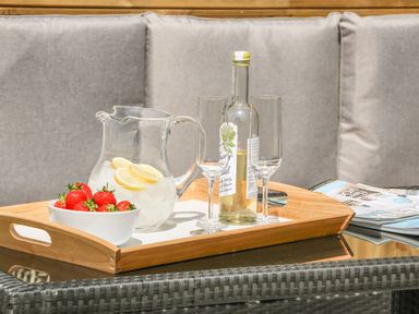 A glass pitcher with lemon slices ice two empty glasses a bottle and a bowl of strawberries on a wooden tray on a wicker table