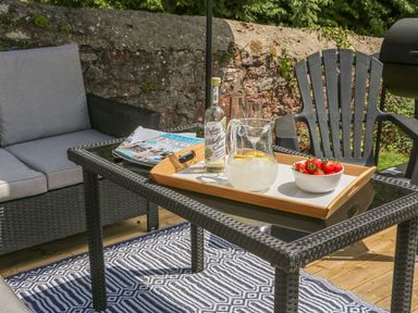 An outdoor seating area with a glass coffee table holding a tray with a bottle, pitcher with ice and lemon, and a bowl of strawberries at The Coach House in Teignmouth